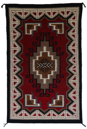 A Teec Nos Pos rug with a Southwestern geometric pattern, featuring a deep red center, beige and brown medallions, and a black and beige border.